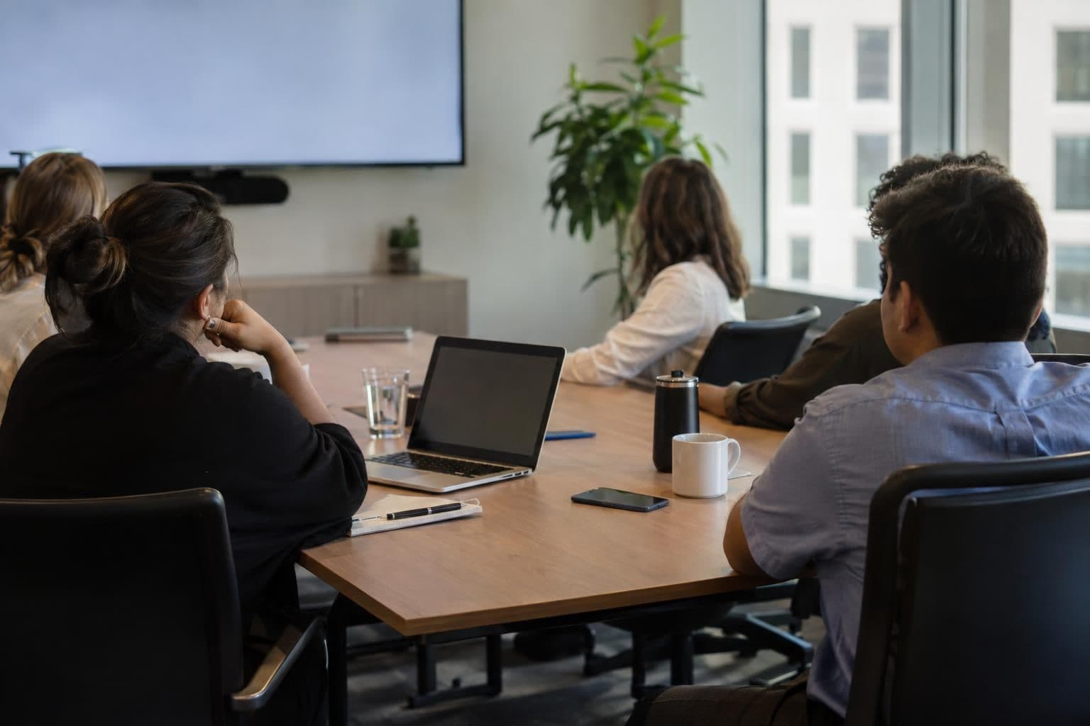 Anonymous attendees sit around a table with a laptop in a plain meeting room.