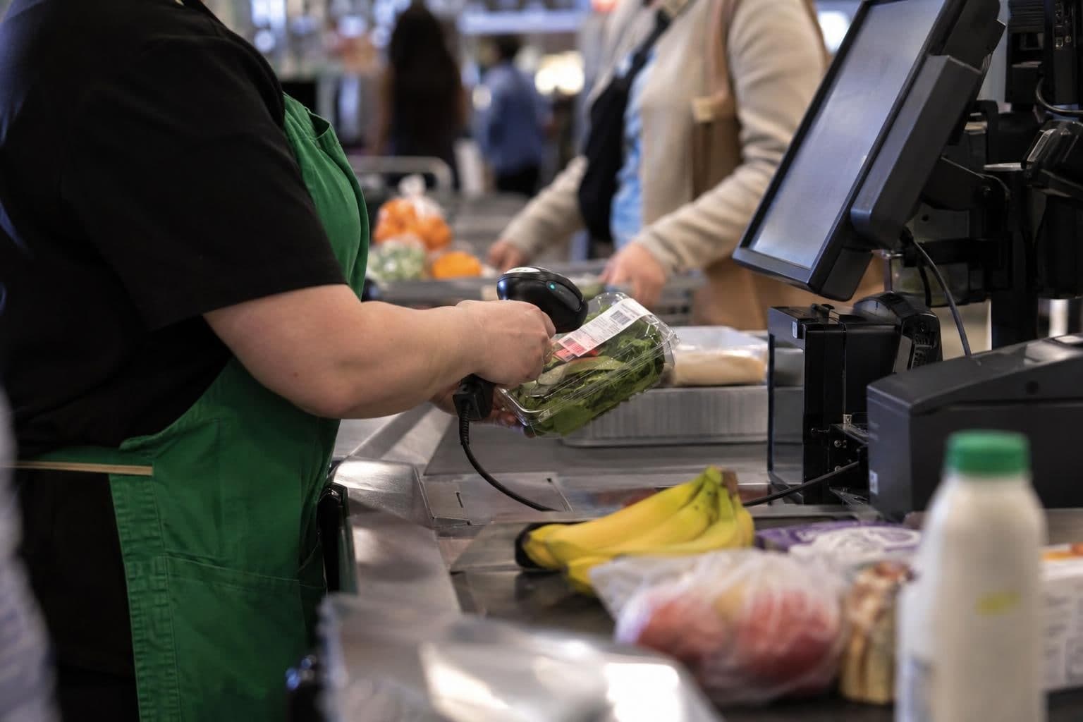 A cashier scans an item at a retail checkout, with shoppers and neighboring counters out of focus.