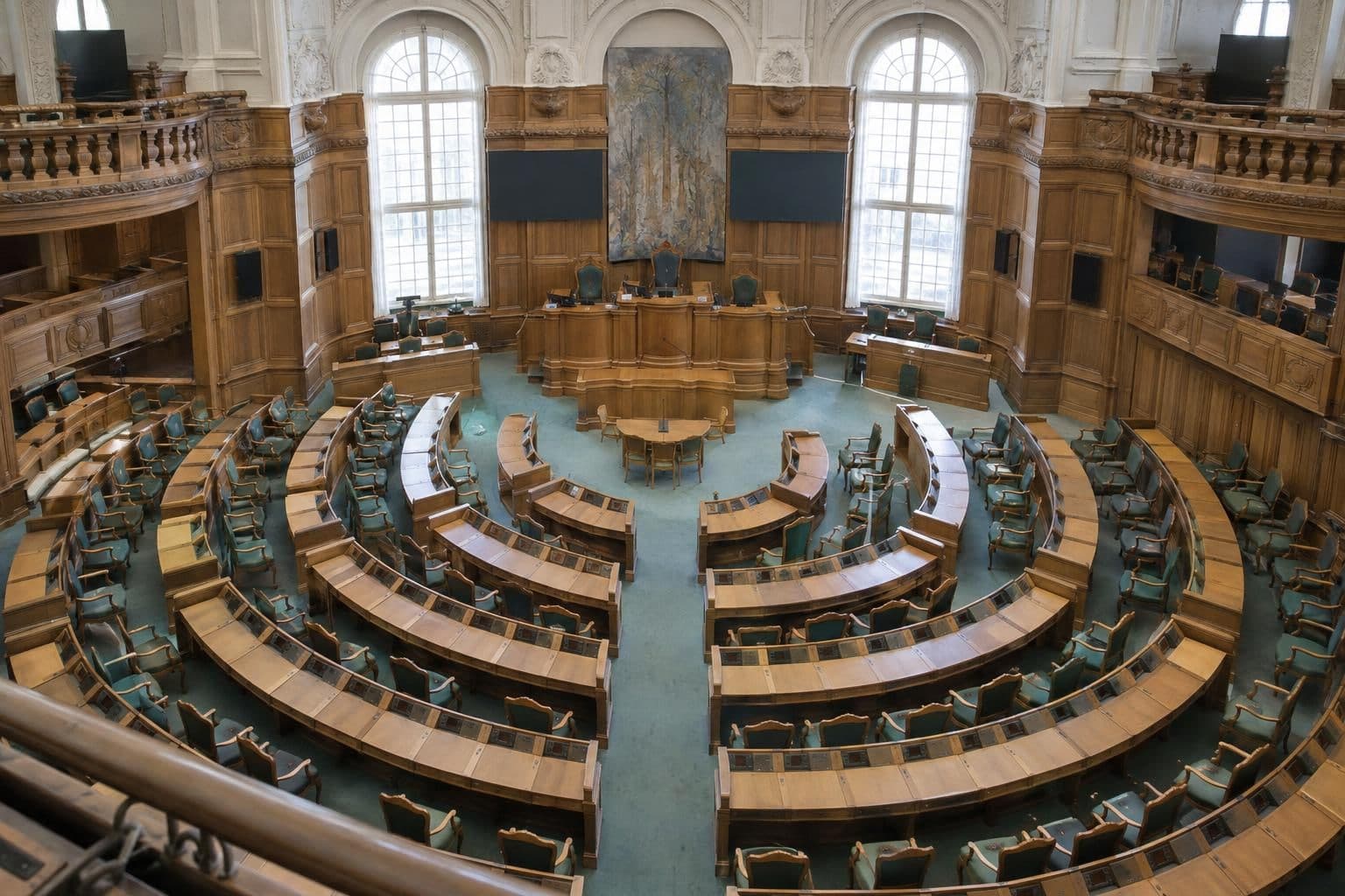 Empty Danish parliament chamber viewed from above, with rows of seats and a central speaking area.