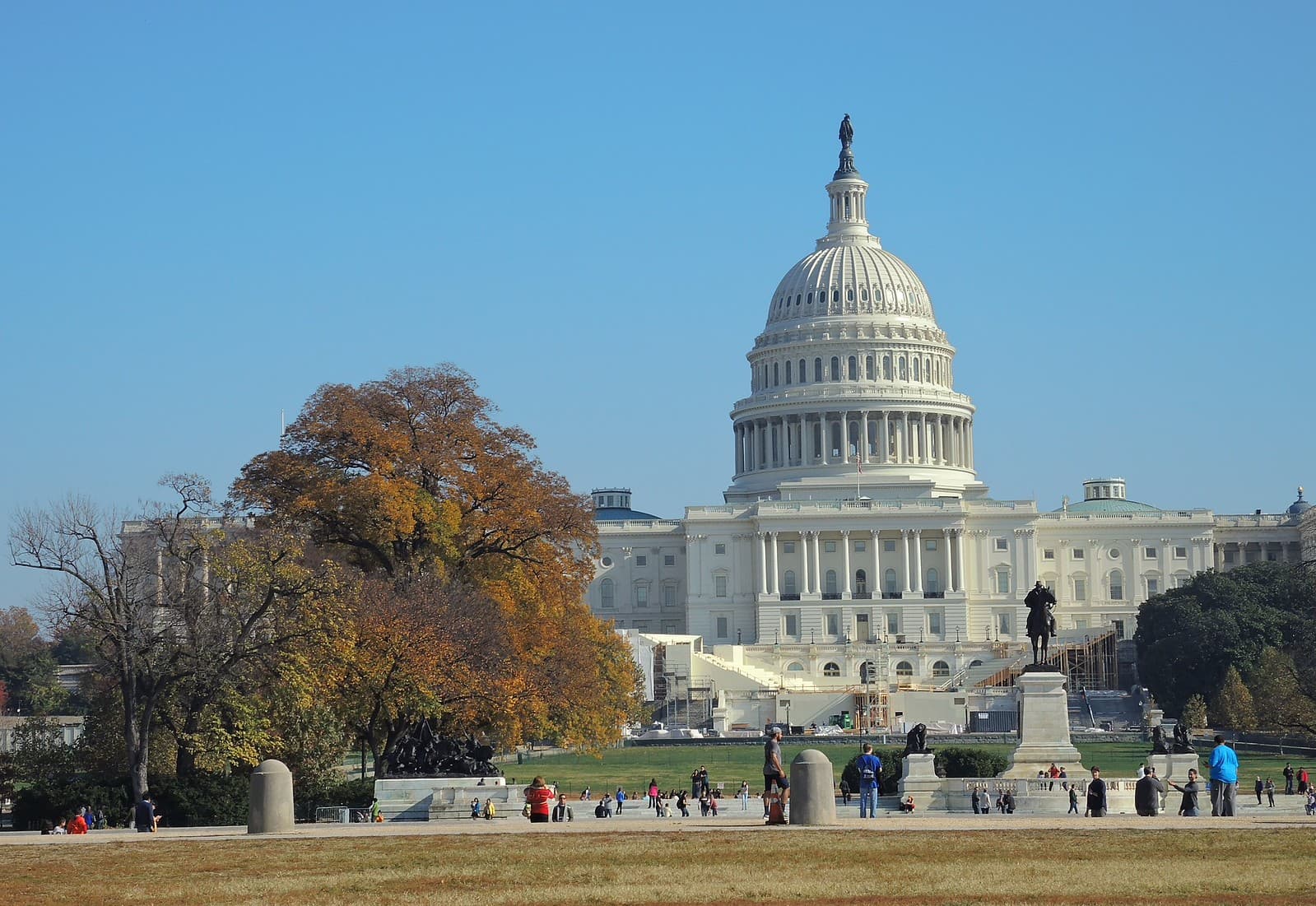 The U.S. Capitol stands beyond its lawn and trees on a clear day, with people walking in the grounds.