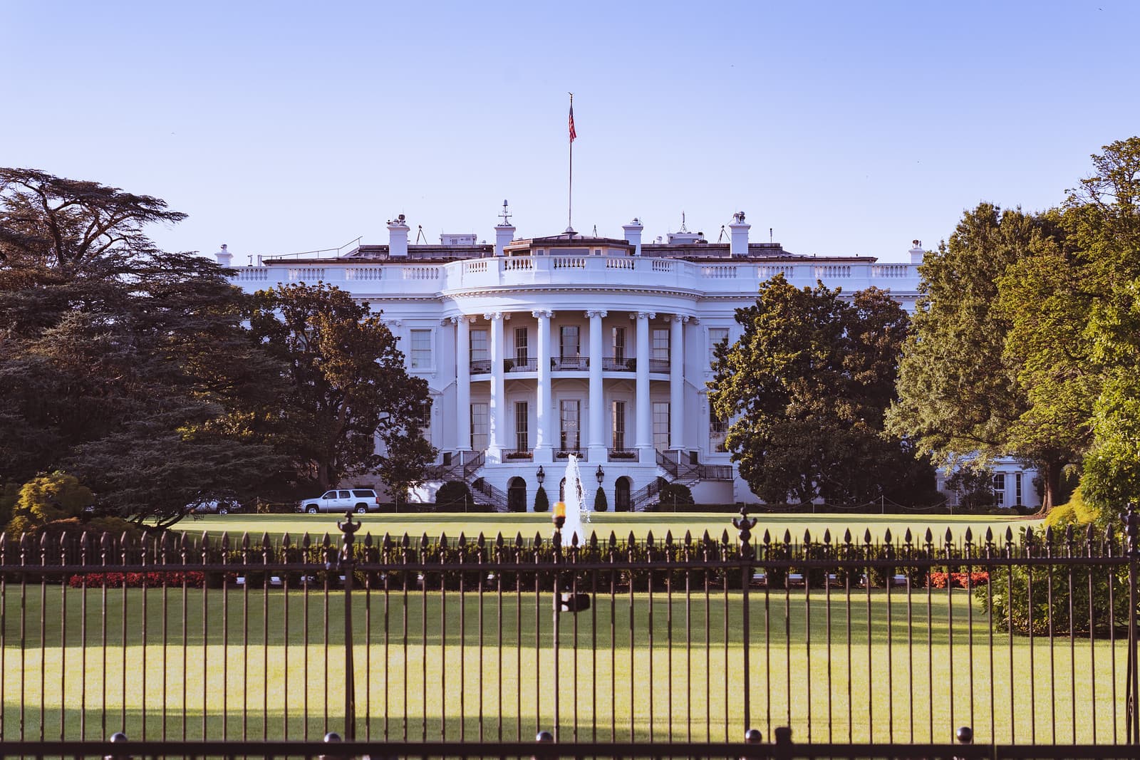 The White House stands beyond a lawn and iron fence, framed by trees.